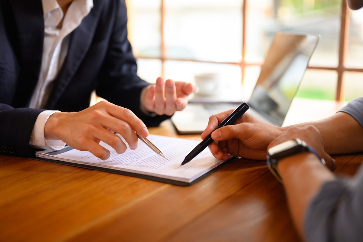 Two people seated at a wooden desk beside a window point pens at a document on a clipboard
