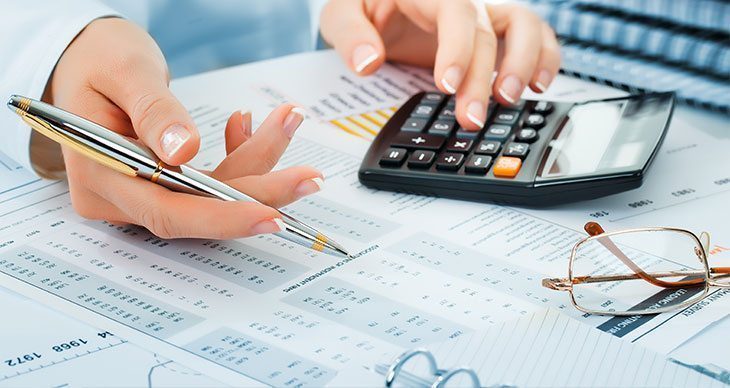 Person using a calculator and holding a pen over financial documents and spreadsheets; a pair of eyeglasses is placed nearby on the desk.