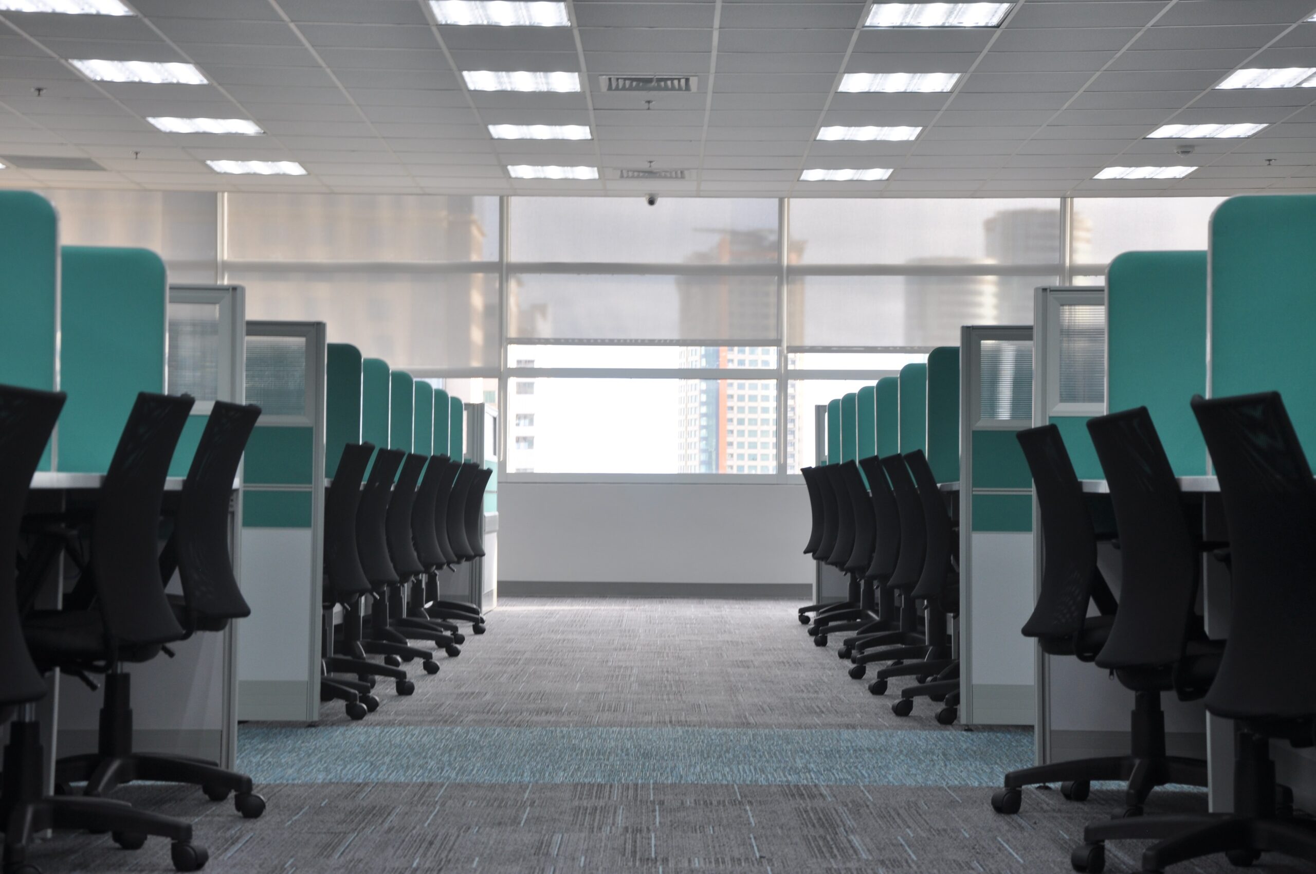 Empty modern office with rows of black chairs and desks separated by teal dividers, facing large windows with city buildings visible outside.