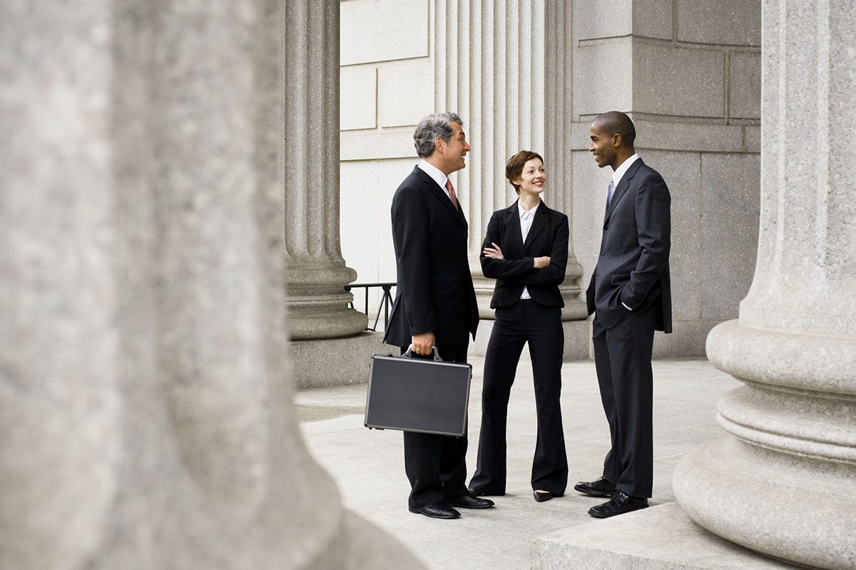 Three businesspeople in suits converse amid stone columns outside of a building