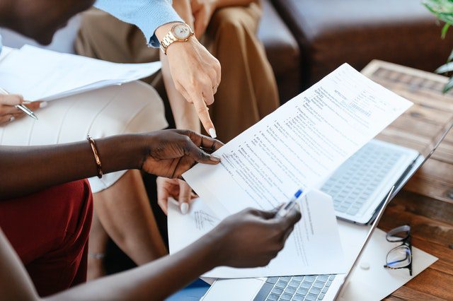 Two people sitting at a table review a printed document together, with laptops, paper, and pens visible on the table. One person is pointing at the document.